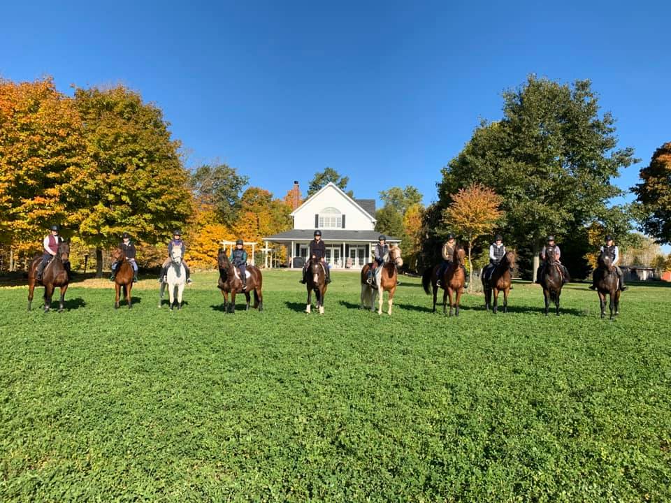 A group of riders on horseback lined up in front of the Sprucehaven Farm house in autumn.